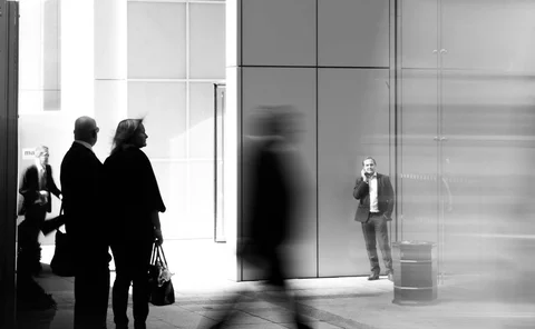 A black and white street photography shot of people outside a modern glass building. A blurred figure walks past in the foreground, while a man in a suit stands in the background talking on a phone.