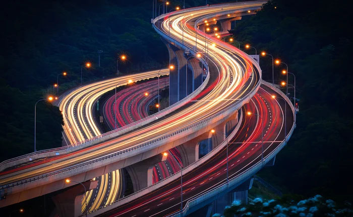 A modern multi-level highway interchange at night, Taipei, featuring winding elevated roads illuminated by vibrant red and golden light trails from moving vehicles, surrounded by dark, forested hills. The intricate infrastructure and flowing traffic trails symbolize connectivity, speed, technological advancement, and efficient transportation networks.