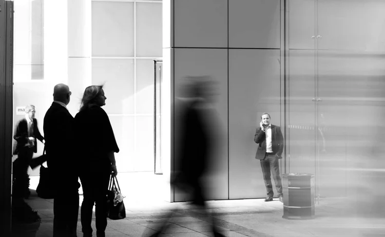 A black and white street photography shot of people outside a modern glass building. A blurred figure walks past in the foreground, while a man in a suit stands in the background talking on a phone.