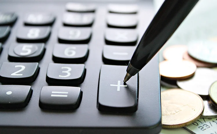 Increased-costs-Getty-958070770 Calculator next to a pile of coins with a pen poised to press the plus key