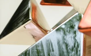 Abstract close-up of overlapping metallic geometric pyramids in green, copper and silver tones, with reflective surfaces against a light background