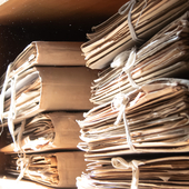 Two piles of dusty beige cardboard files containing paperwork sitting on a shelf with dust motes floating in a shaft of light